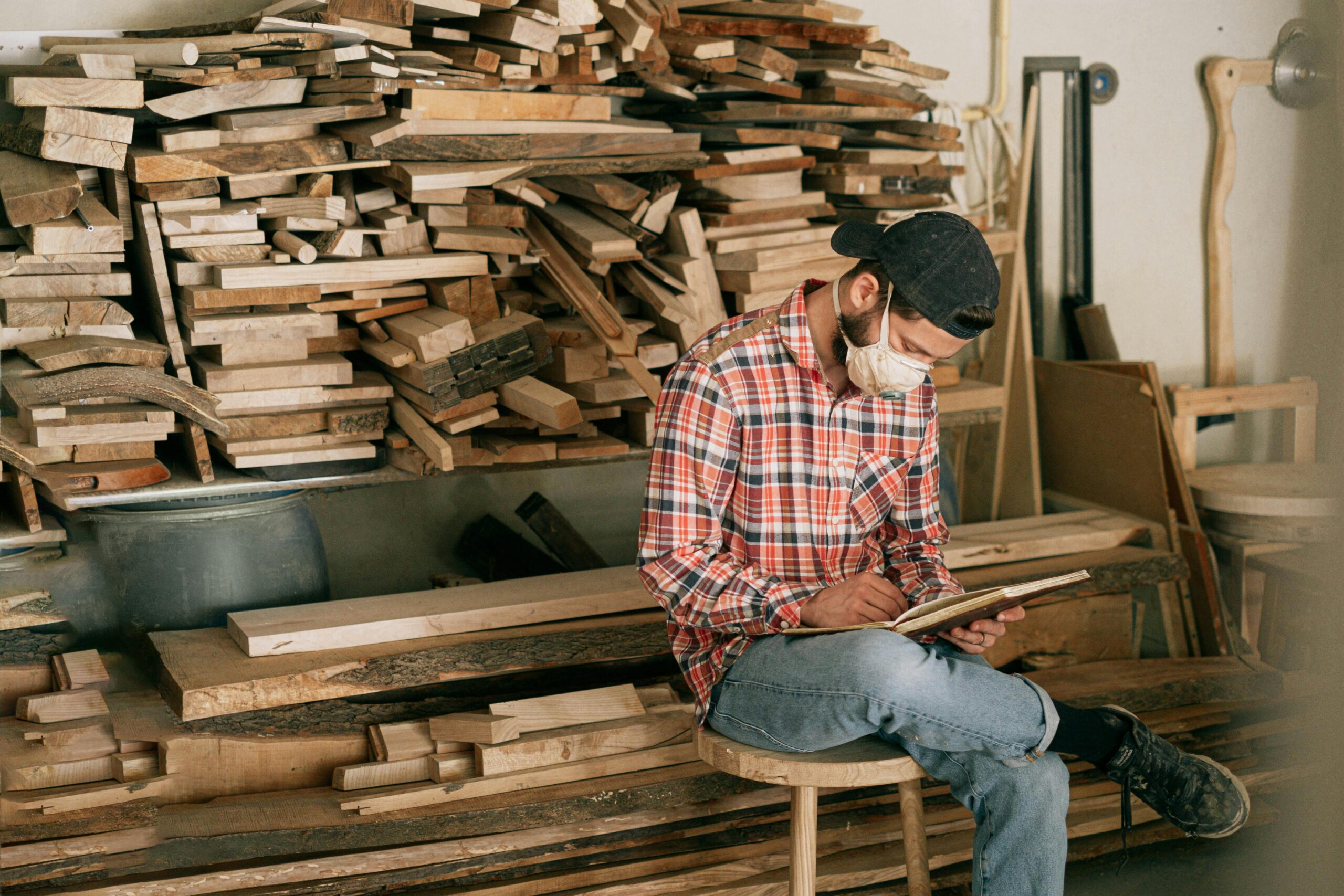 Man wearing mask sketches woodworking designs in a timber-filled workshop.