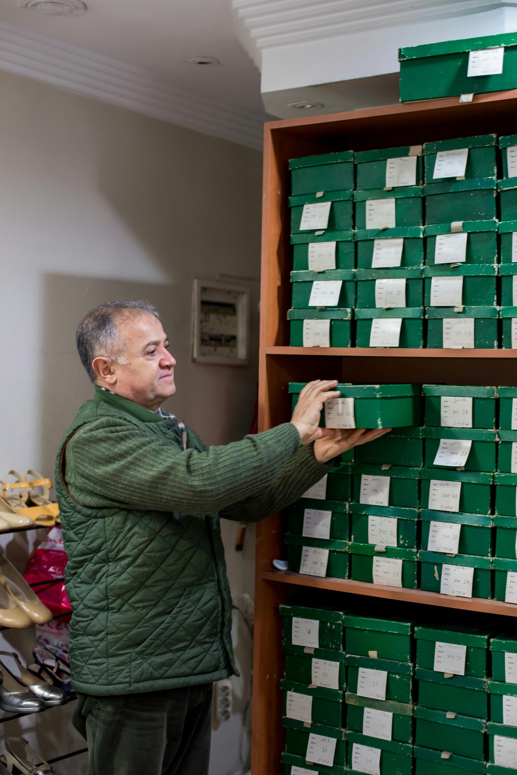 Positive middle aged man in casual outfit putting boxes with shoes on shelves while standing in bright shoe shop near and looking away