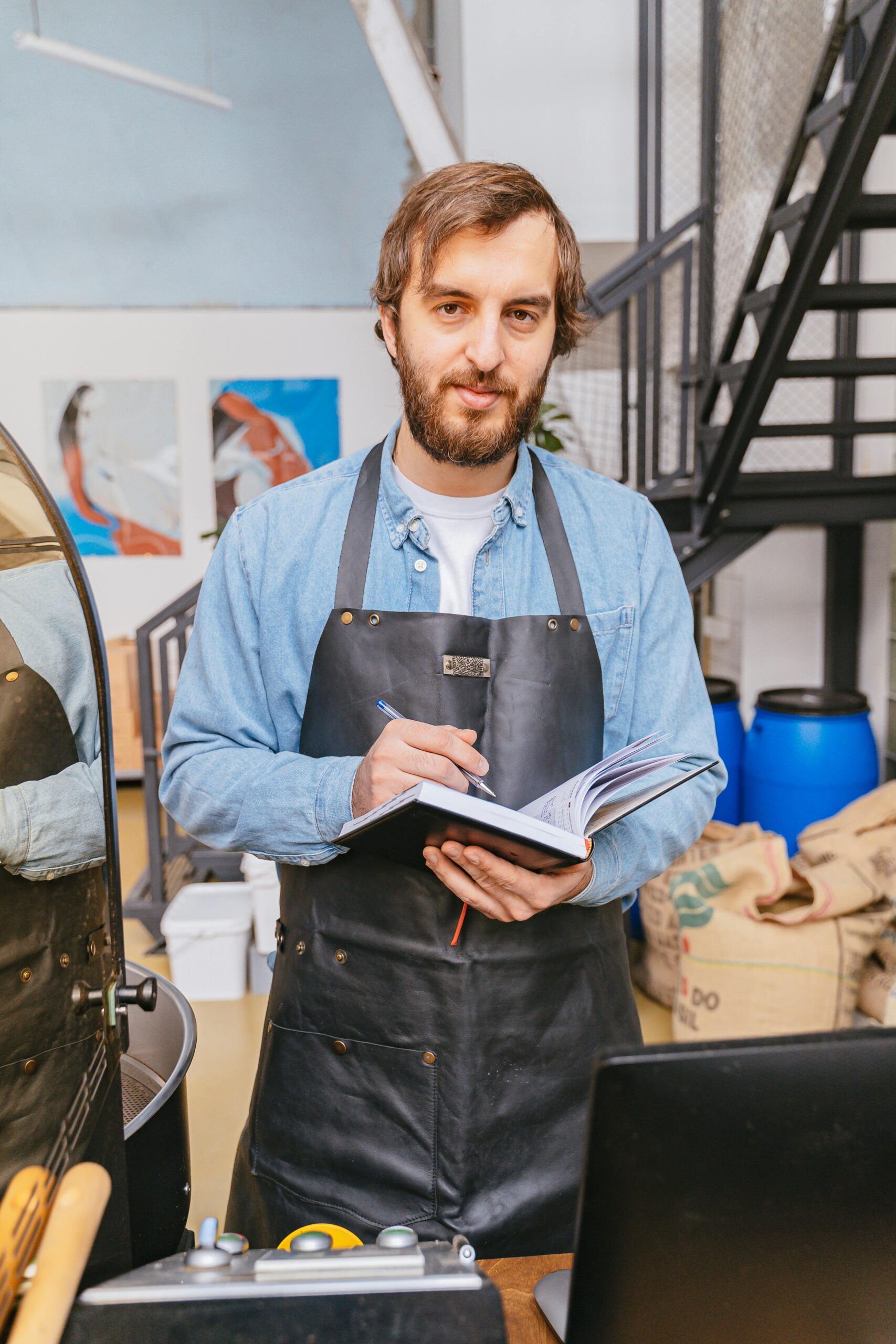 Bearded man in apron holding a notebook and pen inside a coffee shop.
